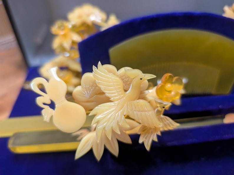 May include: Ivory-coloured hair accessories, including a comb and decorative pieces, are displayed on a blue surface. The comb features a fan shape, while the other pieces showcase intricate bird and floral designs. The accessories are presented in a velvet-lined box.
