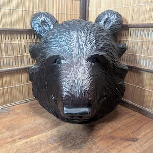 May include: A carved black bear head with detailed fur texture. The bear's face is centered, showcasing its eyes, nose, and ears. The sculpture is placed on a wooden surface, with a bamboo screen in the background.