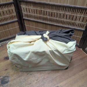 May include: A rectangular box wrapped in a cream-colored cloth, tied with a knot. The top is covered with a dark gray cloth. The box is on a wooden surface, with a bamboo-style wall in the background.