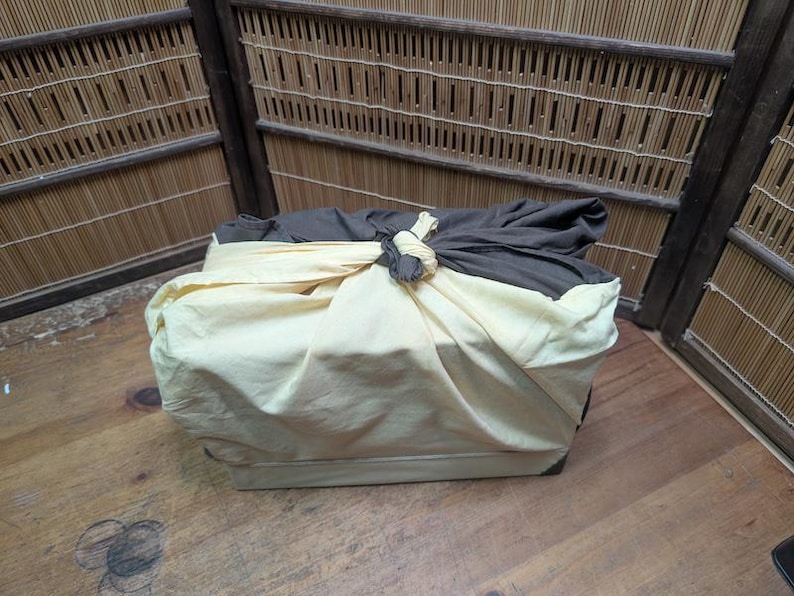 May include: A rectangular box wrapped in a cream-colored cloth, tied with a knot. The top is covered with a dark gray cloth. The box is on a wooden surface, with a bamboo-style wall in the background.