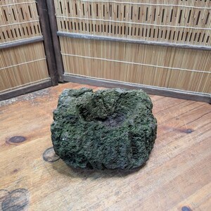 Puede incluir: Un objeto de piedra cuadrado, texturizado y de color verde oscuro, con una depresi&oacute;n central. La piedra tiene una superficie rugosa y porosa y se encuentra sobre una superficie de madera. El fondo presenta una pantalla de bamb&uacute;.