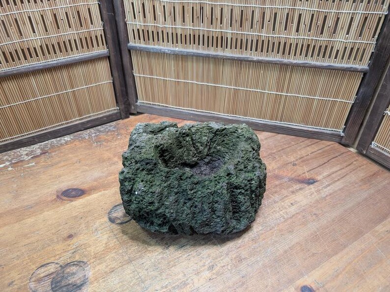 Puede incluir: Un objeto de piedra cuadrado, texturizado y de color verde oscuro, con una depresi&oacute;n central. La piedra tiene una superficie rugosa y porosa y se encuentra sobre una superficie de madera. El fondo presenta una pantalla de bamb&uacute;.