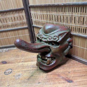 Op de afbeelding: Een gedetailleerd, verweerd, rood en groen Tengu-masker met een lange neus en open mond, die tanden onthult. Het masker staat tegen een houten oppervlak en een bamboe achtergrond, wat traditioneel Japans vakmanschap laat zien.