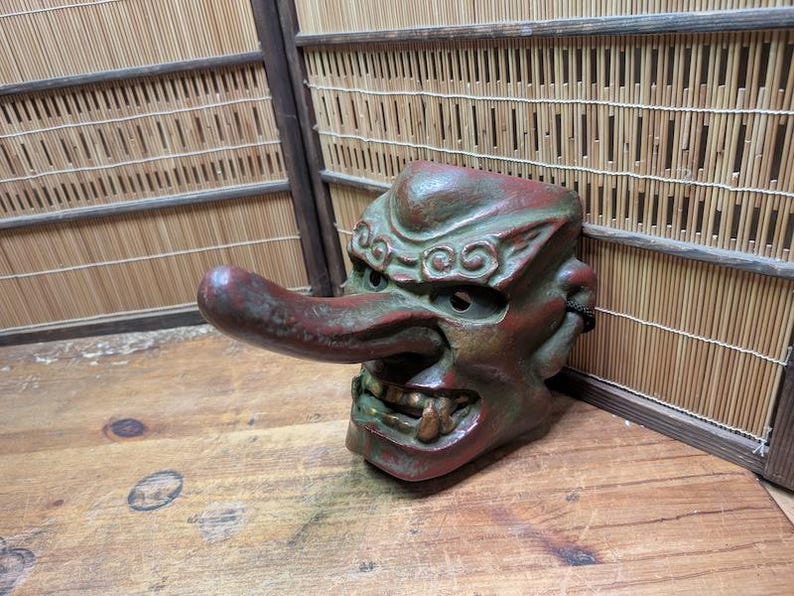 Op de afbeelding: Een gedetailleerd, verweerd, rood en groen Tengu-masker met een lange neus en open mond, die tanden onthult. Het masker staat tegen een houten oppervlak en een bamboe achtergrond, wat traditioneel Japans vakmanschap laat zien.