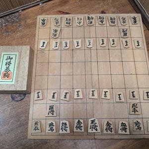 Può includere: Un set da gioco da tavolo Shogi in legno con una scatola di legno. Il tabellone è marrone chiaro con un motivo a griglia e pedine di legno con caratteri giapponesi neri. La scatola ha un'etichetta verde e rossa con testo giapponese.