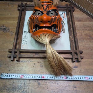May include: An orange and black Japanese Noh mask with a fierce expression, mounted in a wooden frame. The mask has a long, flowing, light brown hairpiece. A ruler is placed below the mask.