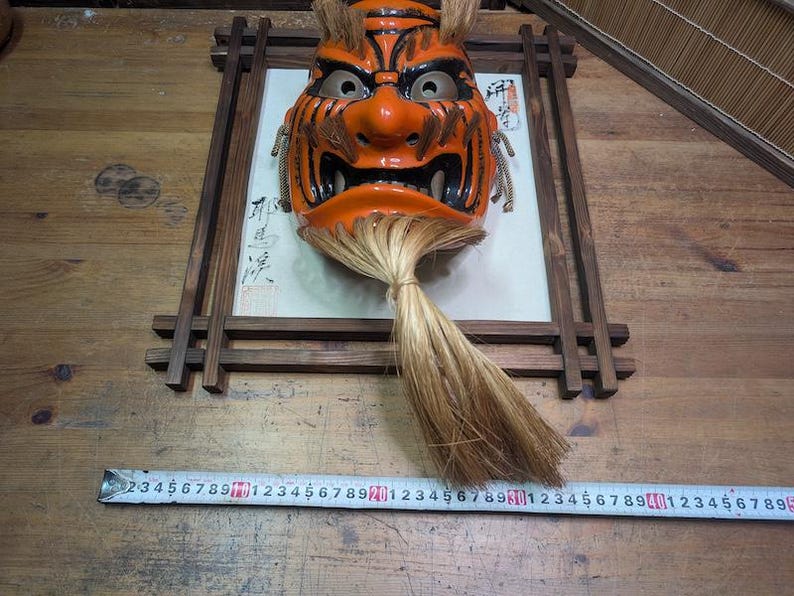 May include: An orange and black Japanese Noh mask with a fierce expression, mounted in a wooden frame. The mask has a long, flowing, light brown hairpiece. A ruler is placed below the mask.
