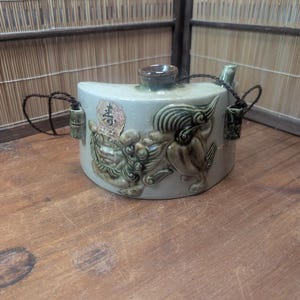 May include: A light green ceramic sake pot with a brown lid and spout. The pot is decorated with a relief of a mythical creature in shades of green and brown. It has two small handles with decorative tassels.