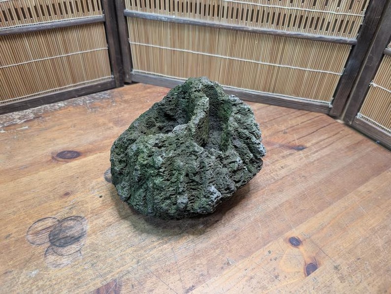 Puede incluir: Una roca texturizada de color verde oscuro con una superficie rugosa descansa sobre una mesa de madera. La roca tiene una forma natural y est&aacute; colocada contra un fondo de madera con un dise&ntilde;o de bamb&uacute;.