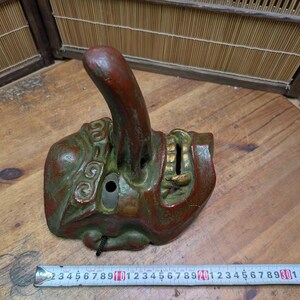 Op de afbeelding: Een antiek Japans Noh-masker, voornamelijk rood en groen met gouden details. Het masker heeft een opvallende, omhoog gebogen neus en een rechthoekige opening. Het masker is ongeveer 13 cm lang. Het masker is verweerd en vertoont tekenen van ouderdom.