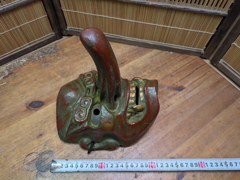 Op de afbeelding: Een antiek Japans Noh-masker, voornamelijk rood en groen met gouden details. Het masker heeft een opvallende, omhoog gebogen neus en een rechthoekige opening. Het masker is ongeveer 13 cm lang. Het masker is verweerd en vertoont tekenen van ouderdom.