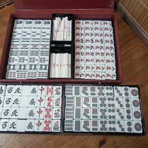 Op de afbeelding: Een complete Mahjong-set in een rode koffer, met tegels gerangschikt in zwarte trays. De tegels zijn wit met zwarte en rode symbolen en karakters. De set bevat rekken voor de tegels en dobbelstenen.