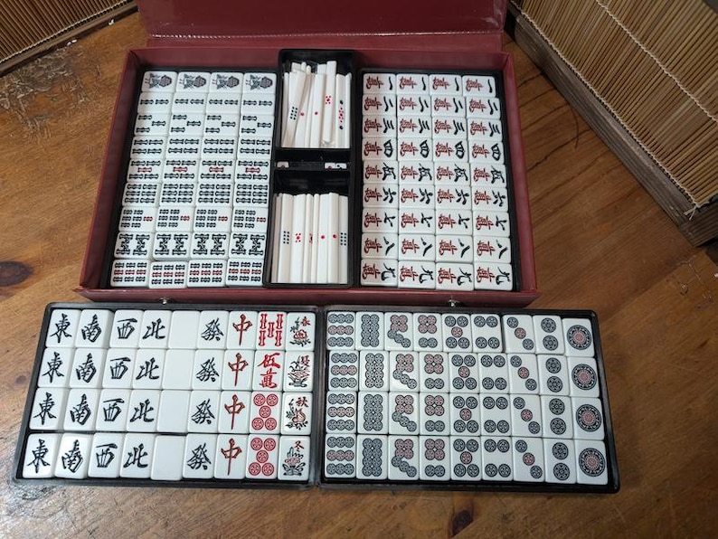 Op de afbeelding: Een complete Mahjong-set in een rode koffer, met tegels gerangschikt in zwarte trays. De tegels zijn wit met zwarte en rode symbolen en karakters. De set bevat rekken voor de tegels en dobbelstenen.