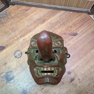 Op de afbeelding: Een vintage Japanse Tengu-masker met een lange neus, rode en groene kleuren en gouden accenten op de tanden. Het masker heeft oogopeningen en is gemaakt van een stevig materiaal. Het is een traditioneel item.