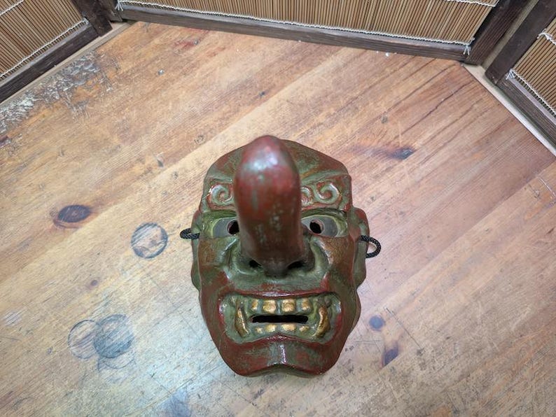 Op de afbeelding: Een vintage Japanse Tengu-masker met een lange neus, rode en groene kleuren en gouden accenten op de tanden. Het masker heeft oogopeningen en is gemaakt van een stevig materiaal. Het is een traditioneel item.