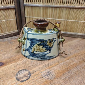 May include: A ceramic sake bottle with a fish design. The bottle is rectangular with a brown spout and handle. The fish is depicted in blue and gold, with a rope handle. The bottle is on a wooden surface.