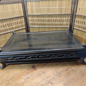 May include: A dark stained wooden display table with carved floral details on the apron. The table has a rectangular top and curved legs. The background includes a bamboo screen.