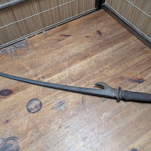 Pode incluir: Uma lâmina de metal curva e antiga com uma guarda em forma de gancho e uma pega enrolada. A lâmina é cinza escuro e a pega é enrolada em um material escuro e texturizado. O objeto está em uma superfície de madeira.