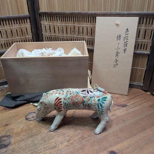 May include: A ceramic pig figurine with floral patterns in red, green, and orange. The figurine is on a wooden surface, next to a wooden box and a cardboard box with Japanese text. The box contains white padding.