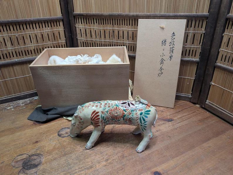May include: A ceramic pig figurine with floral patterns in red, green, and orange. The figurine is on a wooden surface, next to a wooden box and a cardboard box with Japanese text. The box contains white padding.