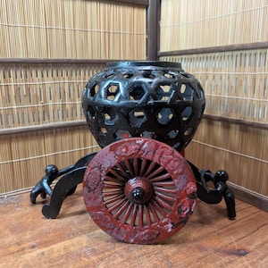 Puede incluir: Un jarrón decorativo negro con un patrón geométrico, apoyado en un soporte negro con una rueda roja. La rueda está intrincadamente tallada con diseños florales. El fondo presenta una pantalla de bambú.