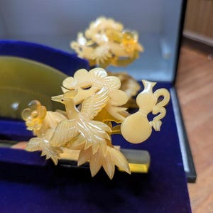May include: A detailed close-up of a decorative hair comb and accessories. The comb and ornaments are carved from a light yellow material, featuring bird and floral designs. The items are displayed on a blue velvet surface.