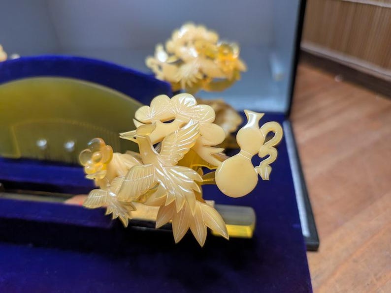 May include: A detailed close-up of a decorative hair comb and accessories. The comb and ornaments are carved from a light yellow material, featuring bird and floral designs. The items are displayed on a blue velvet surface.