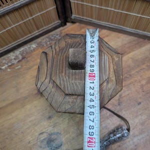May include: A weathered, octagonal wooden object with a square hole in the center. A measuring tape shows the object is approximately 8 inches tall. The wood has visible grain and a natural, aged appearance.