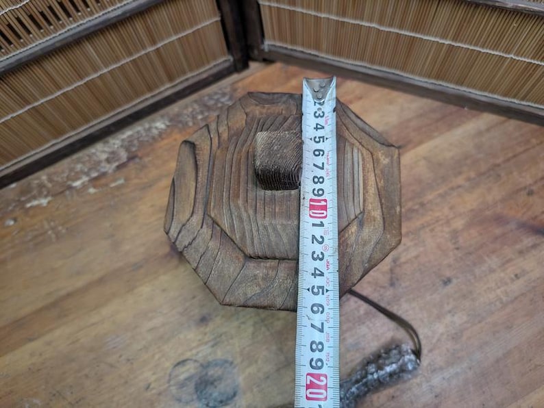 May include: A weathered, octagonal wooden object with a square hole in the center. A measuring tape shows the object is approximately 8 inches tall. The wood has visible grain and a natural, aged appearance.