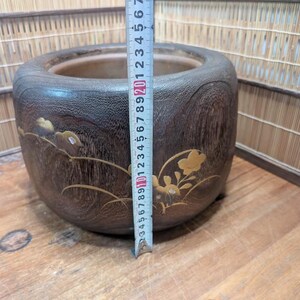 May include: A dark brown wooden planter with a round shape and decorative floral accents. The planter is approximately 6 inches tall and has a 7-inch diameter. The planter is sitting on a wooden surface.