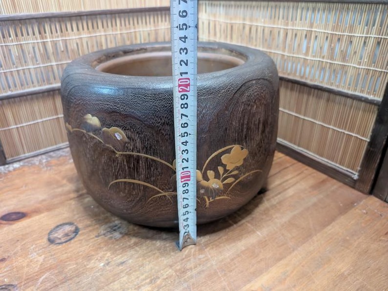 May include: A dark brown wooden planter with a round shape and decorative floral accents. The planter is approximately 6 inches tall and has a 7-inch diameter. The planter is sitting on a wooden surface.