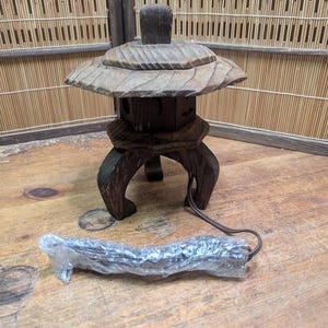 May include: A wooden Japanese garden lantern with a leaf-shaped top and a square body. The lantern has curved legs and a cord. The lantern is brown and sits on a wooden surface.