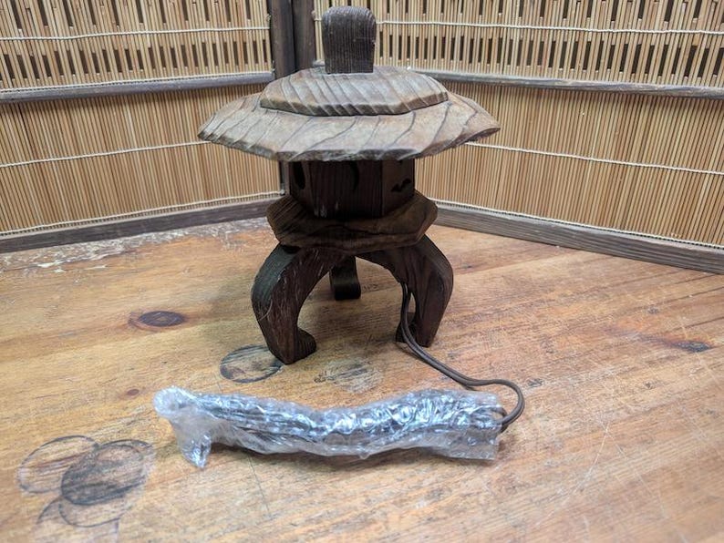 May include: A wooden Japanese garden lantern with a leaf-shaped top and a square body. The lantern has curved legs and a cord. The lantern is brown and sits on a wooden surface.
