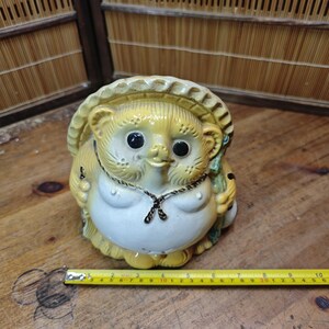 May include: A ceramic tanuki statue with a straw hat, a white belly, and black eyes. The statue has a necklace and is holding a small object. A yellow measuring tape is next to the statue.