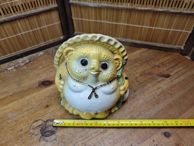 May include: A ceramic tanuki statue with a straw hat, a white belly, and black eyes. The statue has a necklace and is holding a small object. A yellow measuring tape is next to the statue.