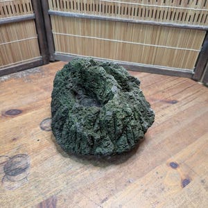 Puede incluir: Una roca verde oscura y texturizada con una superficie rugosa y una depresi&oacute;n central. La roca est&aacute; sobre una superficie de madera, con una pantalla de bamb&uacute; en el fondo. La roca parece ser un elemento decorativo.