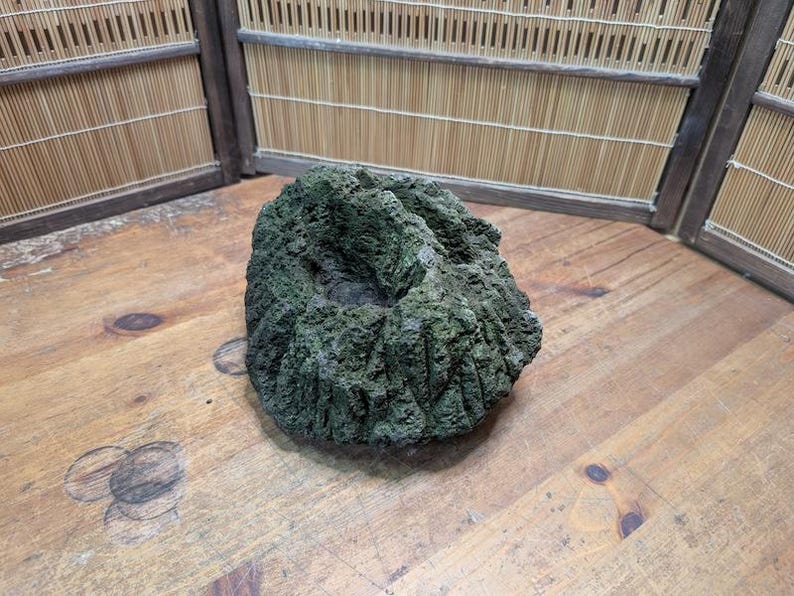Puede incluir: Una roca verde oscura y texturizada con una superficie rugosa y una depresi&oacute;n central. La roca est&aacute; sobre una superficie de madera, con una pantalla de bamb&uacute; en el fondo. La roca parece ser un elemento decorativo.