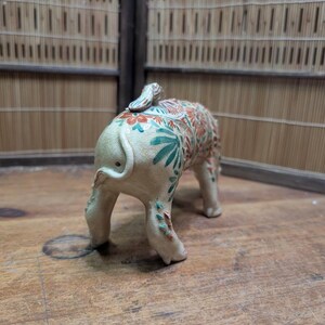 May include: A ceramic elephant figurine with a cream-colored base and intricate floral patterns in shades of orange, green, and blue. The elephant is standing on four legs, with its tail curled up. The background is a wooden surface.