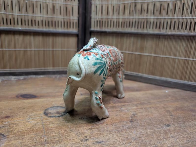 May include: A ceramic elephant figurine with a cream-colored base and intricate floral patterns in shades of orange, green, and blue. The elephant is standing on four legs, with its tail curled up. The background is a wooden surface.