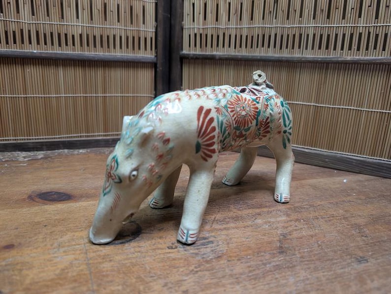 May include: A ceramic figurine of an animal with a long snout, possibly a tapir or similar creature. The figurine is cream-colored with red, green, and blue floral patterns. A small figure sits on its back.