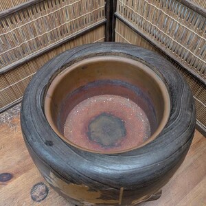 May include: A large, round, dark-toned ceramic pot with a wide rim and a textured, wood-like exterior. The interior reveals a reddish-brown hue. The pot sits on a wooden surface, with a bamboo-style backdrop.