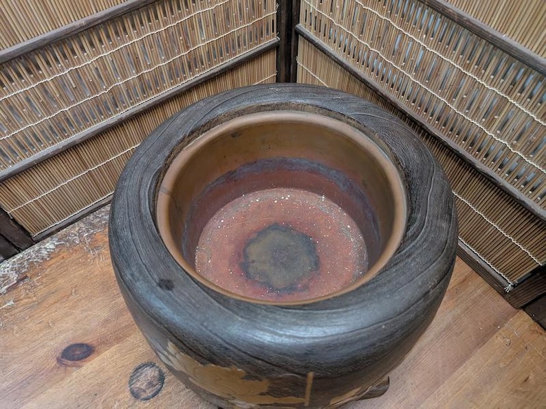May include: A large, round, dark-toned ceramic pot with a wide rim and a textured, wood-like exterior. The interior reveals a reddish-brown hue. The pot sits on a wooden surface, with a bamboo-style backdrop.