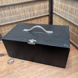 May include: A black wooden box with a silver handle and clasp. The box is rectangular and appears to be made of wood. The handle is arched, and the clasp is decorative. The box is sitting on a wooden surface.