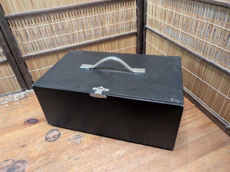 May include: A black wooden box with a silver handle and clasp. The box is rectangular and appears to be made of wood. The handle is arched, and the clasp is decorative. The box is sitting on a wooden surface.