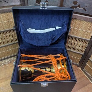 K&ouml;nnte beinhalten: Ein schwarzer rechteckiger Koffer mit blauem Samtinterieur, der eine traditionelle japanische Trommel enth&auml;lt. Die Trommel ist schwarz mit goldenen Akzenten und orangefarbenem Seil. Der Koffer ist ge&ouml;ffnet und zeigt die Trommel.
