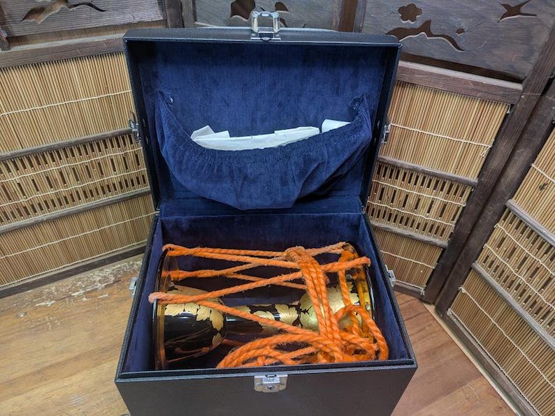 K&ouml;nnte beinhalten: Ein schwarzer rechteckiger Koffer mit blauem Samtinterieur, der eine traditionelle japanische Trommel enth&auml;lt. Die Trommel ist schwarz mit goldenen Akzenten und orangefarbenem Seil. Der Koffer ist ge&ouml;ffnet und zeigt die Trommel.