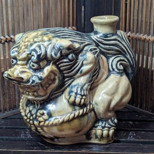 May include: A ceramic sculpture of a mythical creature, featuring a lion-like body with intricate detailing. The piece is primarily beige with blue and brown accents, and a small spout on the back. The sculpture is placed on a dark wooden surface.