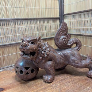 May include: A brown, carved statue of a mythical creature, possibly a guardian lion, holding a decorative ball. The statue has a detailed mane, open mouth, and a stylized tail. The ball has cut-out shapes. The statue is on a wooden surface.