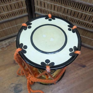 K&ouml;nnte beinhalten: Kleine dekorative Trommel mit cremefarbenem Deckel mit schwarzem Rand und floralen Akzenten. Die Trommel hat ein orangefarbenes Seil und einen goldfarbenen K&ouml;rper. Der Hintergrund umfasst eine Holzoberfl&auml;che und einen gewebten Bambus-Schirm.
