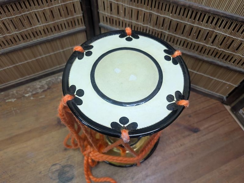 K&ouml;nnte beinhalten: Kleine dekorative Trommel mit cremefarbenem Deckel mit schwarzem Rand und floralen Akzenten. Die Trommel hat ein orangefarbenes Seil und einen goldfarbenen K&ouml;rper. Der Hintergrund umfasst eine Holzoberfl&auml;che und einen gewebten Bambus-Schirm.